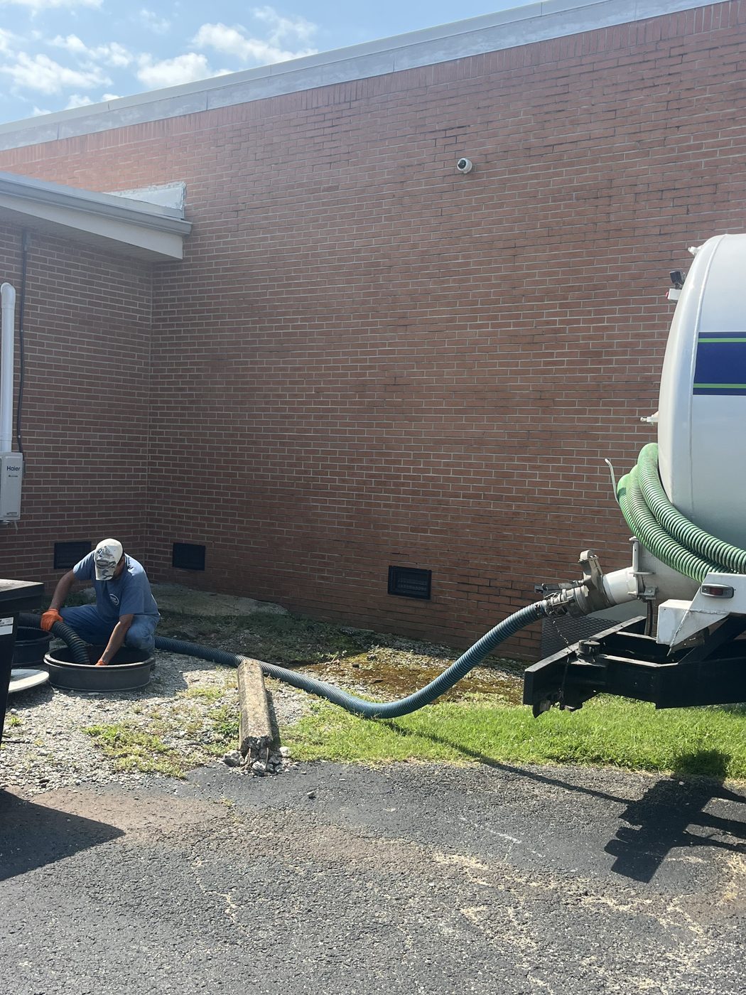 Drain Works technician pumping an outdoor grease trap beside a commercial brick building with a vacuum hose connected to the pump truck.