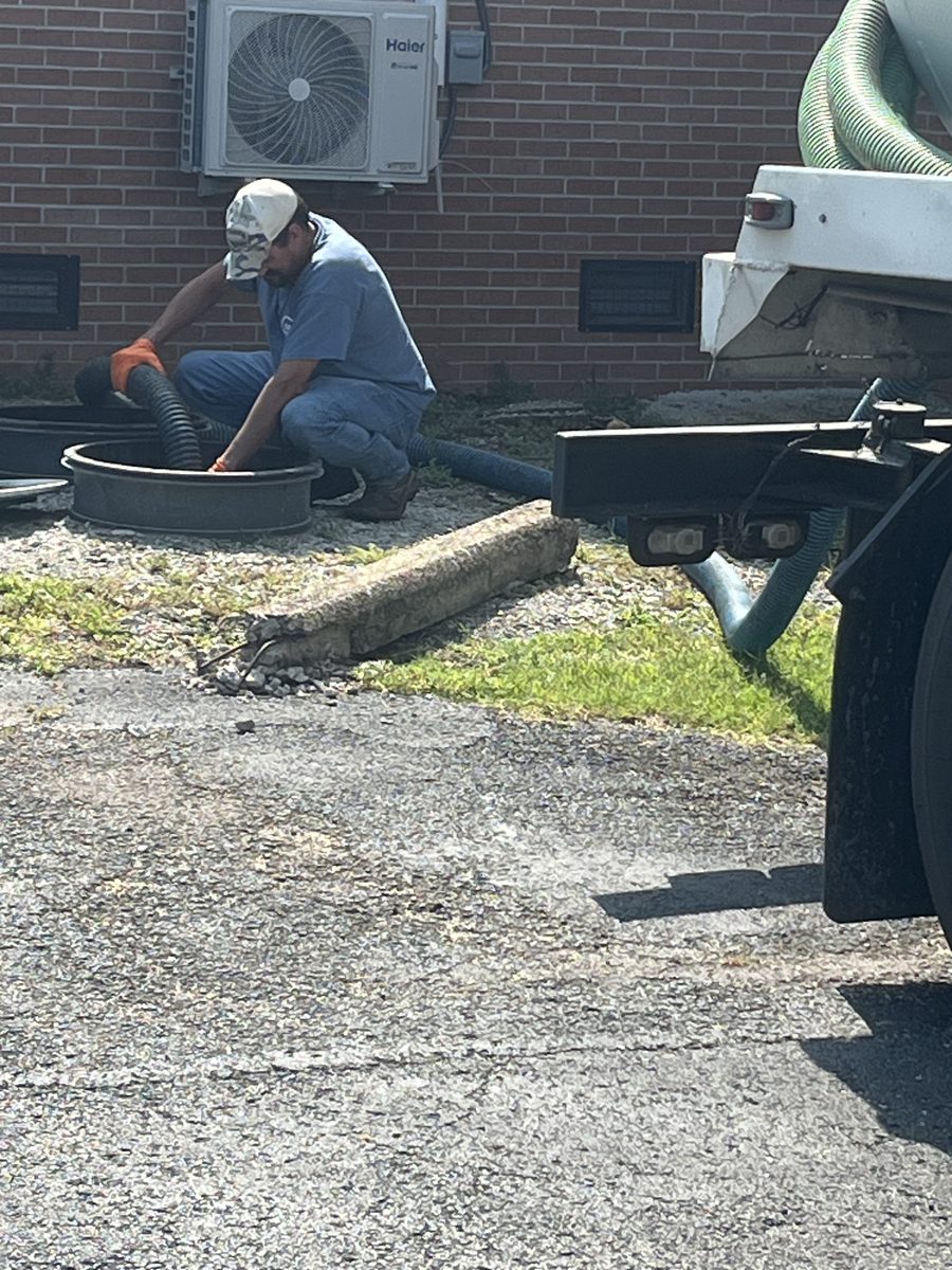 Close-up of a technician wearing gloves operating a vacuum hose at an outdoor grease trap access point beside a pump truck.