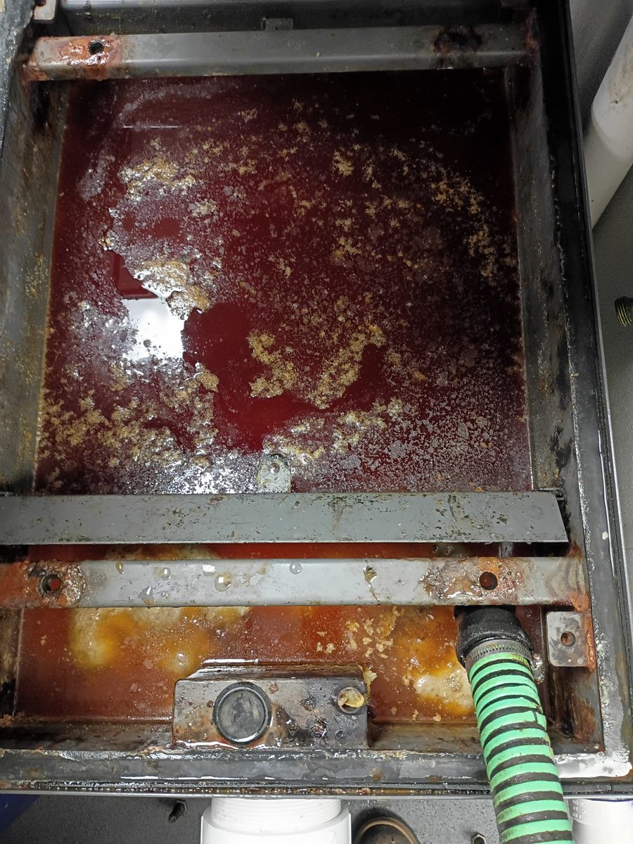 Before grease trap cleaning, an indoor grease trap compartment is filled with brown wastewater, floating grease solids, and residue around the metal edges.